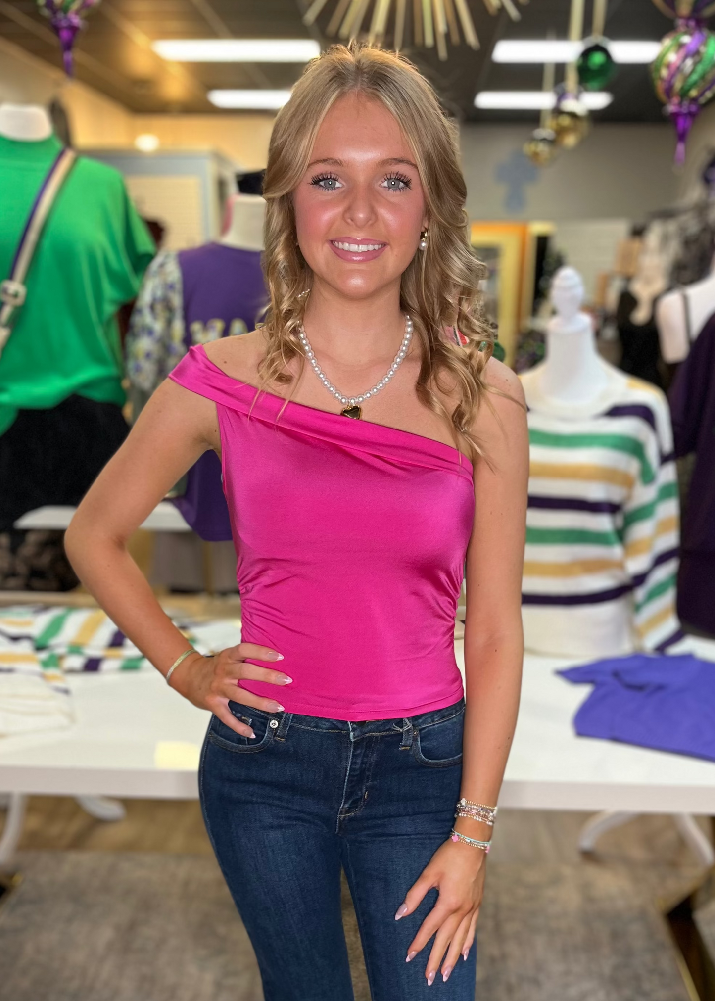 A smiling blonde tries our soft cotton candy-pink one-shoulder tank and dark jeans among sweet displays. Elle Boutique, Zachary, LA.