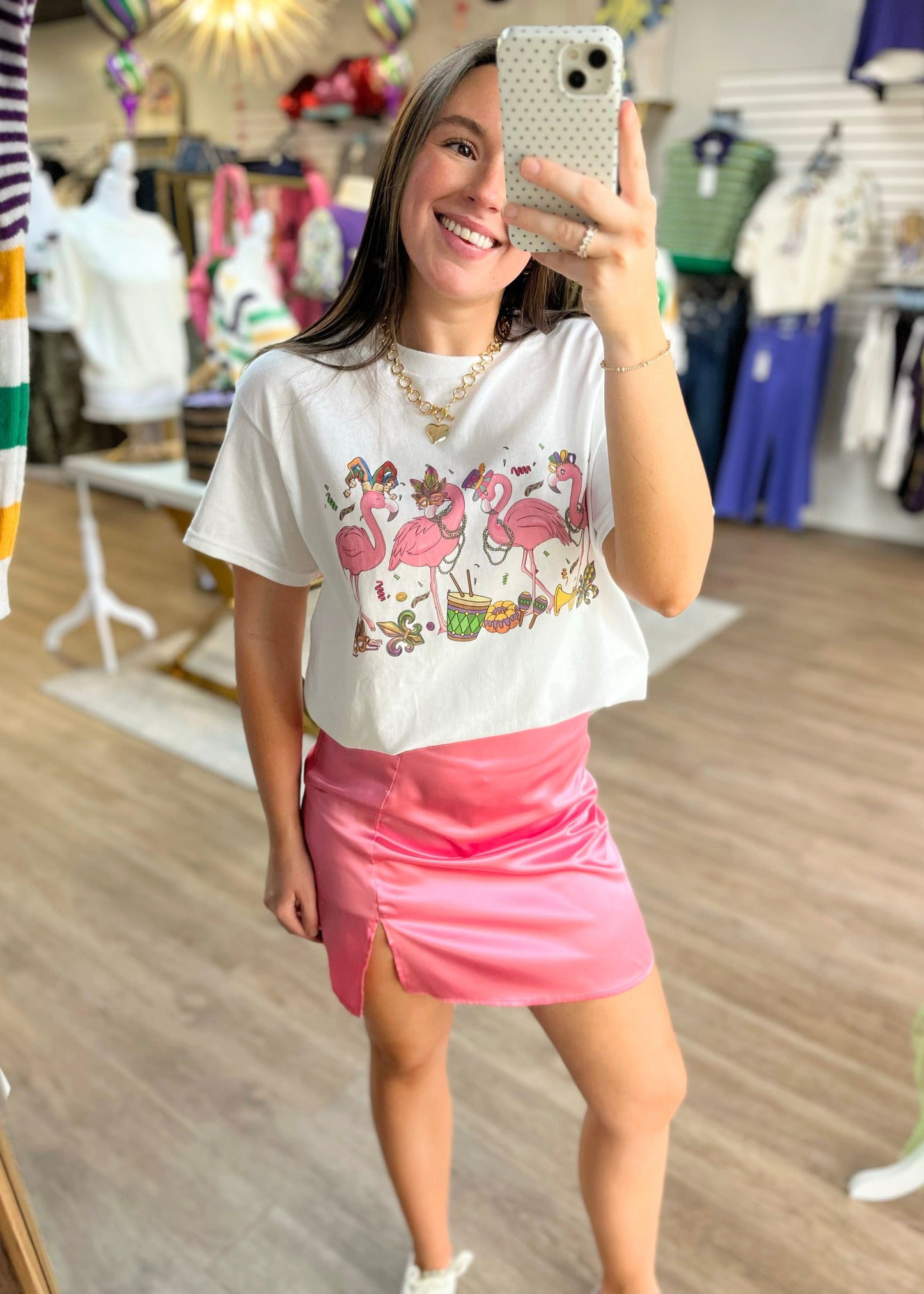 A cheerful gal snaps a mirror selfie in a pink satin mini skirt and soft flamingo tee, surrounded by cute finds. Elle Boutique, Zachary, LA.