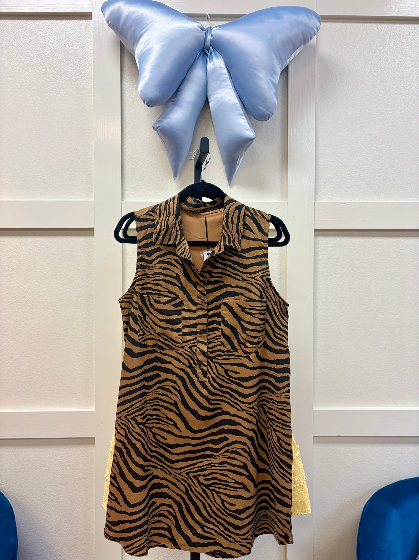 Our Tiger Takeover Pocket Dress is the perfect choice for easygoing days, featuring a breezy sleeveless pullover fit and soft, swishy fabric with playful brown and black tiger stripes. Handy side pockets make it as practical as it is cute. Shown here on a hanger beneath a pretty blue satin butterfly against classic white paneled walls—this one’s ready to bring some wild charm to your wardrobe! Elle Boutique, Zachary, LA.