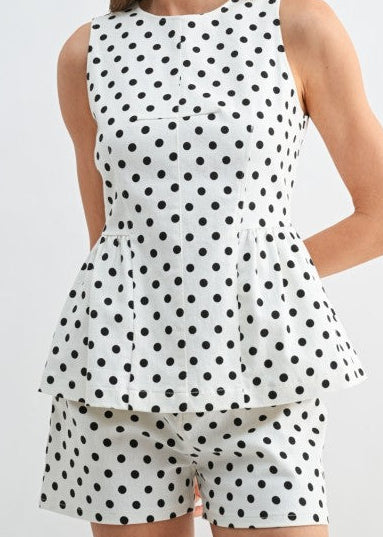 A smiling woman stands against a soft light background, showing off our Dot Your I’s Twill Shorts—the sweetest cotton-blend, mid-rise shorts sprinkled with playful polka dots. She’s paired them with a breezy sleeveless white peplum top, creating a charming Southern look that’s comfy and chic for warm weather days. Elle Boutique, Zachary, LA.
