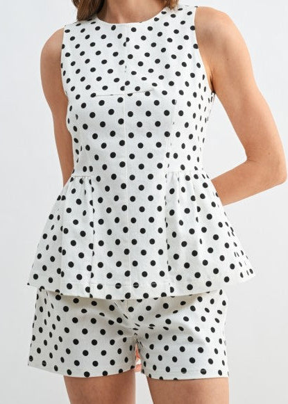 Our model shows off the charming “Dot Your I’s Peplum Top,” a sleeveless ivory blouse sprinkled with classic black polka dots, featuring a sweet peplum hem for that extra twirl. The soft, lightweight twill fabric keeps things cool and comfy, while the matching shorts create an effortlessly put-together look. Perfect for sunny days and casual gatherings, set against a simple light backdrop. Elle Boutique, Zachary, LA.