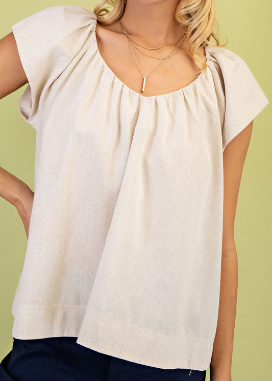 Soft linen top in light beige with short sleeves, a gathered neckline, and dark pants. Gold necklaces add charm. Elle Boutique, Zachary, LA.