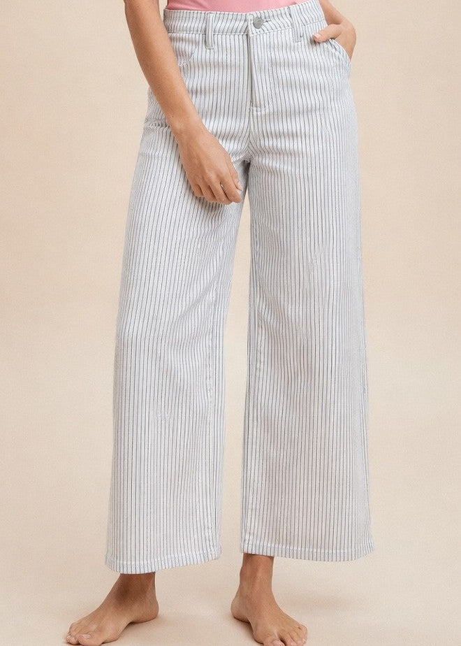 A lovely lady stands barefoot against a soft beige background, wearing our Preppy Stripe - White & Denim Pinstripe Jeans. These jeans are made from cozy stretch cotton and feature delicate vertical blue stripes for that classic Southern charm. She pairs them with a sweet pink top and tucks one hand casually in her pocket, showing off the relaxed fit that’s perfect for sunny days. Elle Boutique, Zachary, LA.