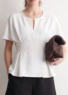 A lovely lady styles our cream Perfectly Pleated Peplum Top, featuring soft, flowy fabric and darling short sleeves, paired with classic black pants. The gentle pleats give a relaxed yet flattering fit that’s easy to dress up or down. She carries a rich dark brown clutch, all set against a simple light background—just right for Southern days when comfort meets charm. Elle Boutique, Zachary, LA.