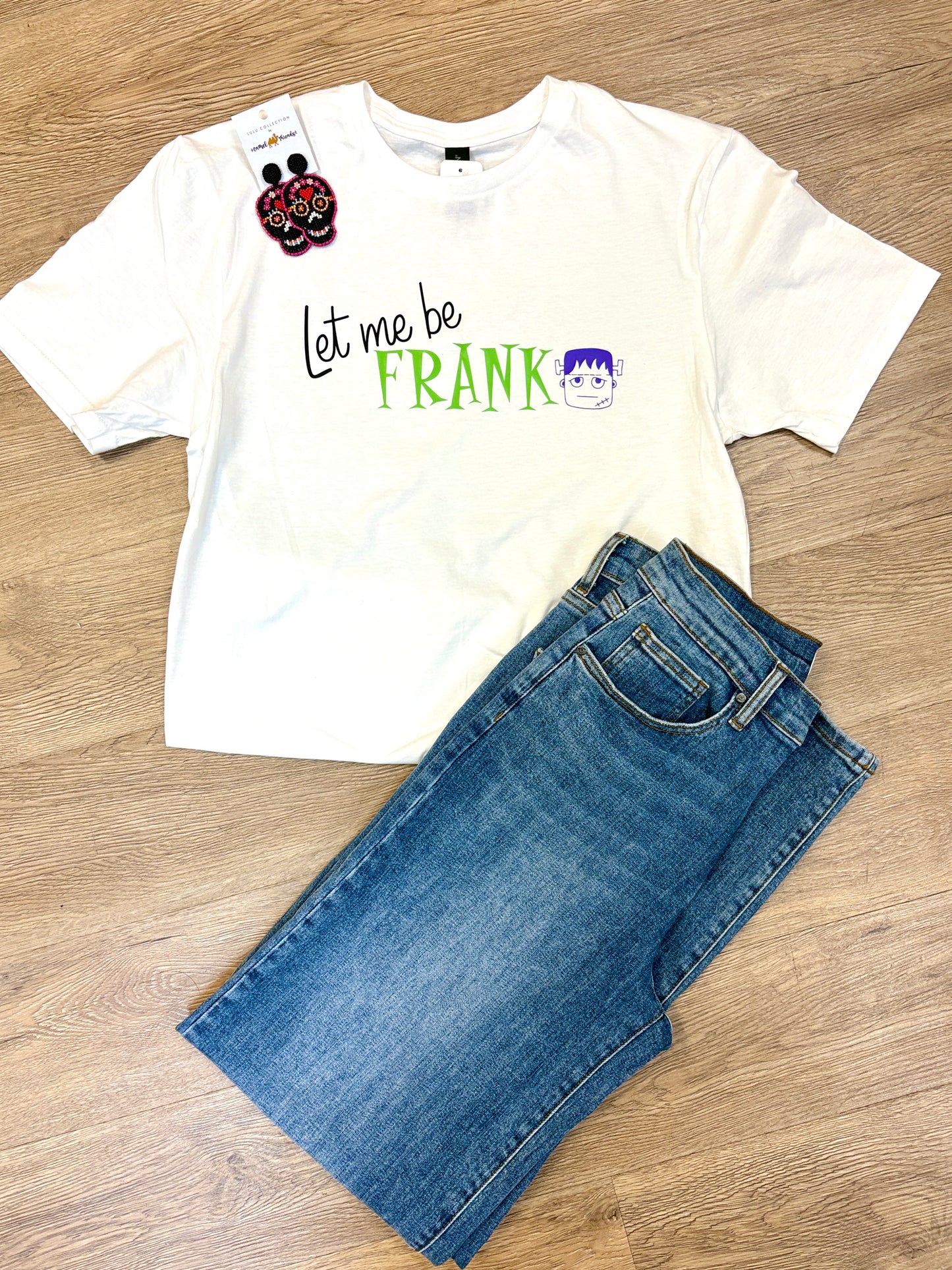 This comfy white cotton tee adds a playful touch to your day with its cheerful “Let me be FRANK” message and a cute cartoon Frankenstein face across the front. Styled here with classic blue jeans and festive sugar skull earrings for a laid-back Southern vibe, it’s perfect for relaxed weekends or casual fun. The soft fabric and easy fit keep you cozy from morning coffee to evening strolls. Pictured on a warm wood floor at Elle Boutique, Zachary, LA.