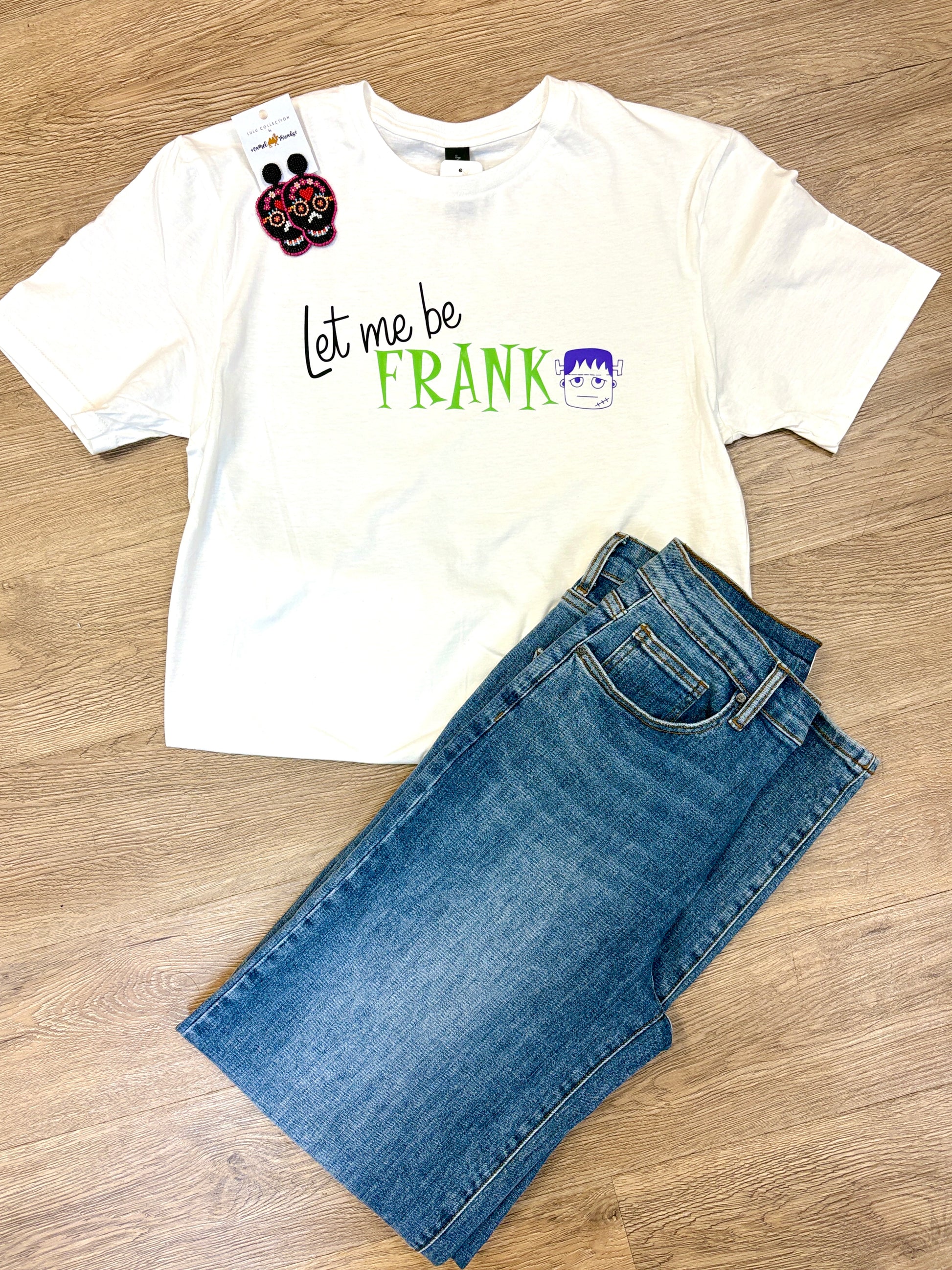 This comfy white cotton tee adds a playful touch to your day with its cheerful “Let me be FRANK” message and a cute cartoon Frankenstein face across the front. Styled here with classic blue jeans and festive sugar skull earrings for a laid-back Southern vibe, it’s perfect for relaxed weekends or casual fun. The soft fabric and easy fit keep you cozy from morning coffee to evening strolls. Pictured on a warm wood floor at Elle Boutique, Zachary, LA.
