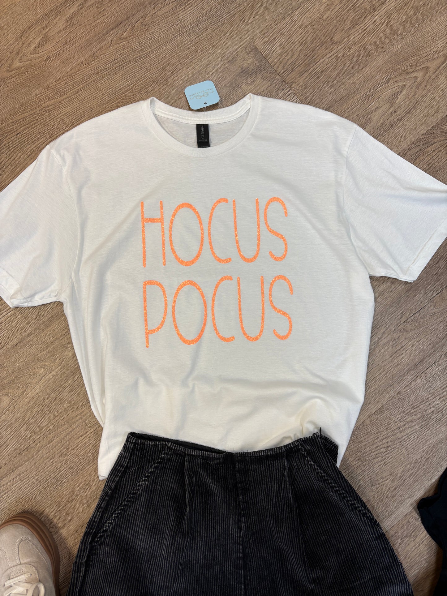 Snuggle into this soft, comfy white cotton tee featuring playful orange “HOCUS POCUS” lettering and a touch of Southern charm. Shown here laid flat on a rustic wooden floor next to dark jeans and the edge of your favorite casual shoe, its relaxed fit makes it perfect for those easygoing fall days or cozy get-togethers with friends. Ideal for ladies who adore a little magic in their wardrobe. Elle Boutique, Zachary, LA.