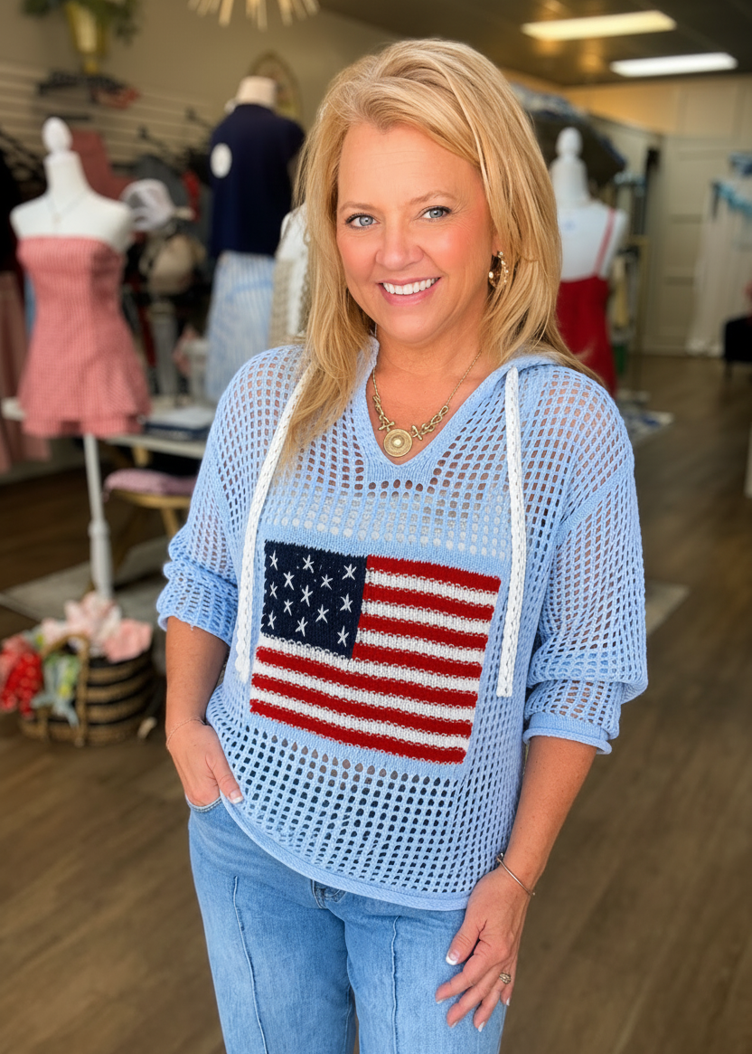 A cheerful blonde models a light blue crochet hoodie with an American flag knit, paired with denim. Elle Boutique, Zachary, LA.