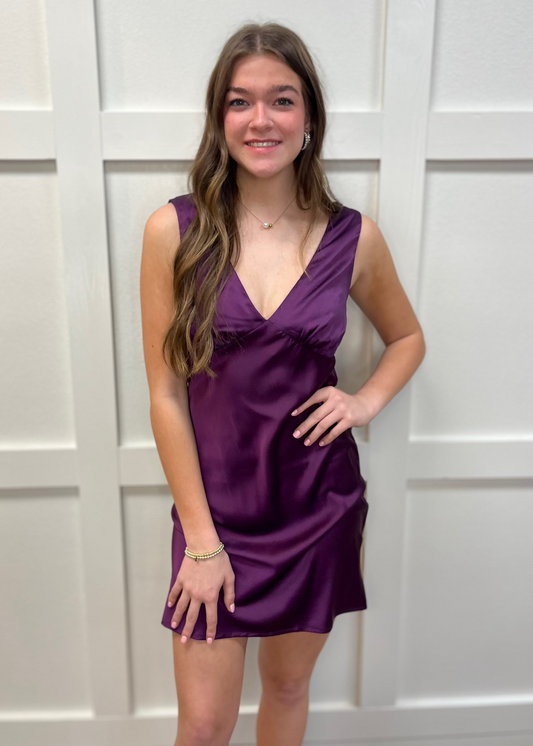 A young woman in a flowy plum satin sleeveless dress smiles sweetly, styled with dainty jewelry. Elle Boutique, Zachary, LA.