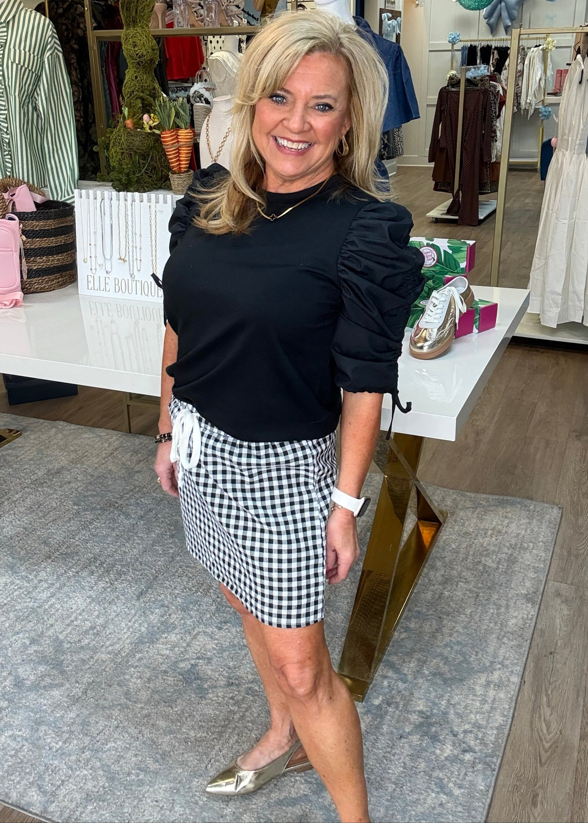 A smiling woman in a soft cotton black & white gingham skirt with drawstring waist, black puff-sleeve top, silver shoes. Elle Boutique, Zachary, LA.