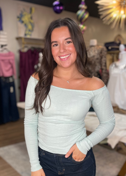 A smiling young woman wears a mint green off-shoulder cotton-blend top with a relaxed fit and dark jeans at Elle Boutique, Zachary, LA.