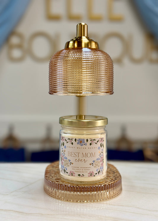 A cozy “Best Mom Ever” candle sits under a Sweet Water Decor lamp warmer, glowing softly by blue drapes. Elle Boutique, Zachary, LA.