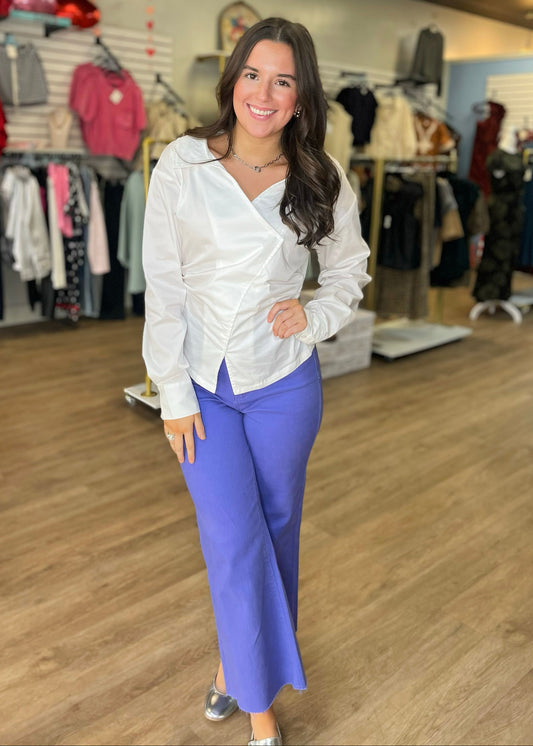 A lady beams in a cozy shop, wearing a crisp cotton blouse, silver shoes, and bright blue stretch high-rise wide-leg denim. Elle Boutique, Zachary, LA.