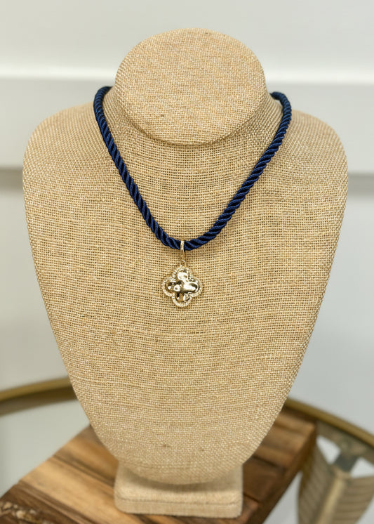 Gold clover pendant with pretty details hangs on a chunky navy rope, shown on burlap. Elle Boutique, Zachary, LA.