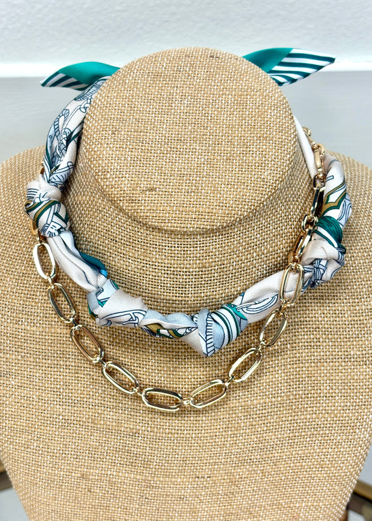 A bust displays our Seaside Nights Chain Necklace, soft twisted fabric in green, white, gold and gold chain, tied in a sweet bow. Elle Boutique, Zachary, LA.
