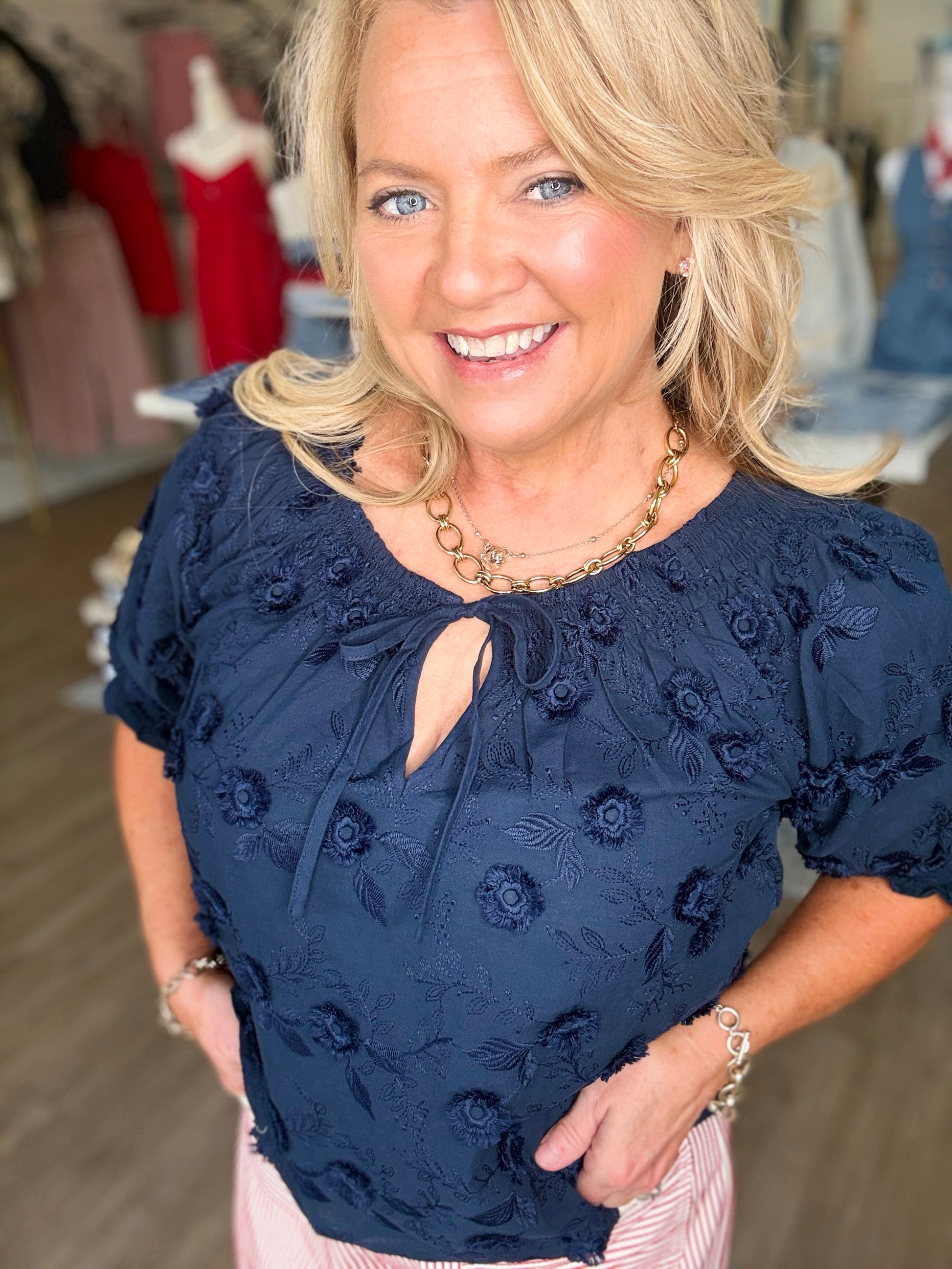 A smiling blonde shows off the Midnight Magnolia Top—soft cotton, relaxed fit, elegant embroidery—in Elle Boutique, Zachary, LA.