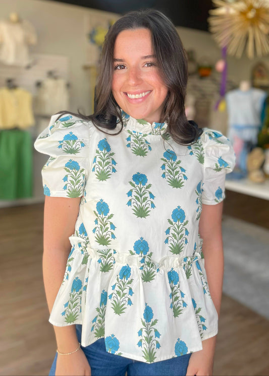 A smiling woman wears our soft cotton Blue Bloom Peplum Top—blue-green florals, ruffled sleeves & waist. Elle Boutique, Zachary, LA.