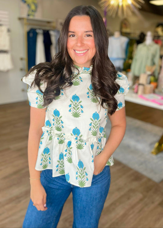 A woman with long brown hair beams in a boutique, wearing our cotton Blue Bloom Peplum Top with flutter sleeves and jeans. Elle Boutique, Zachary, LA.