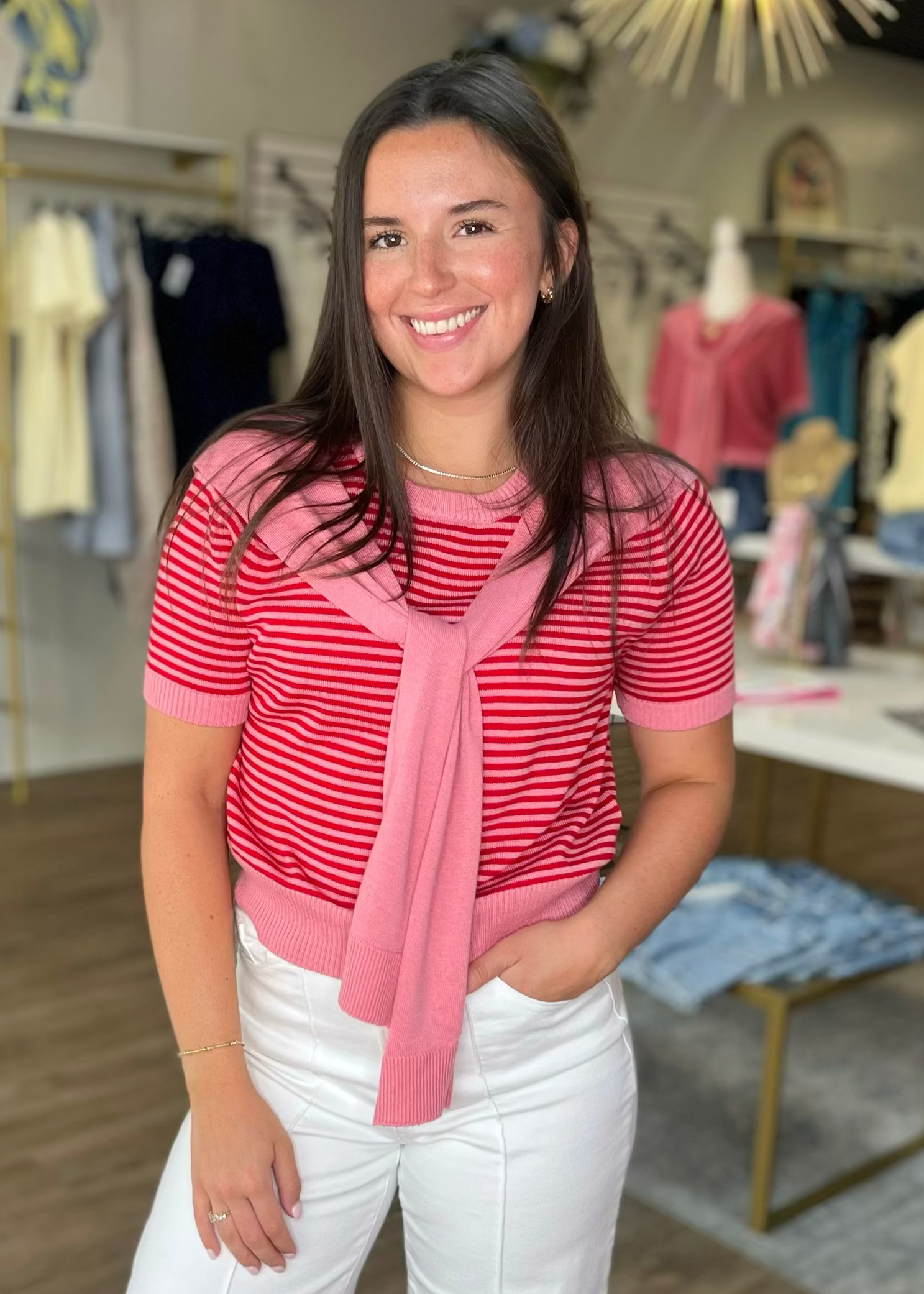 A smiling woman wears The Paris Tie Knit Top in soft cotton, topped with a pink sweater and white pants. Elle Boutique, Zachary, LA.