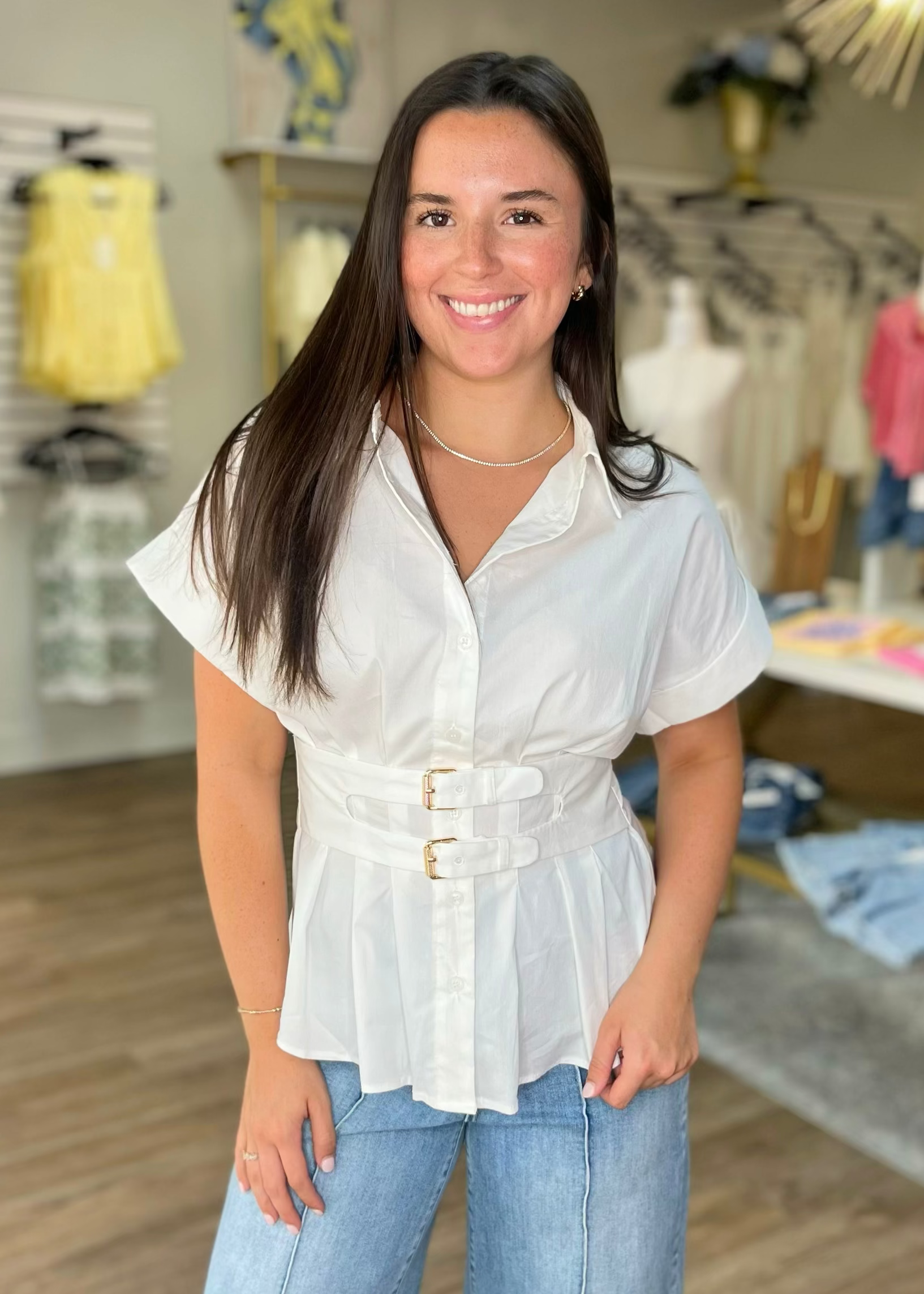 A woman with long brown hair smiles in a boutique, wearing a crisp white belted peplum top and denim jeans. Elle Boutique, Zachary, LA.