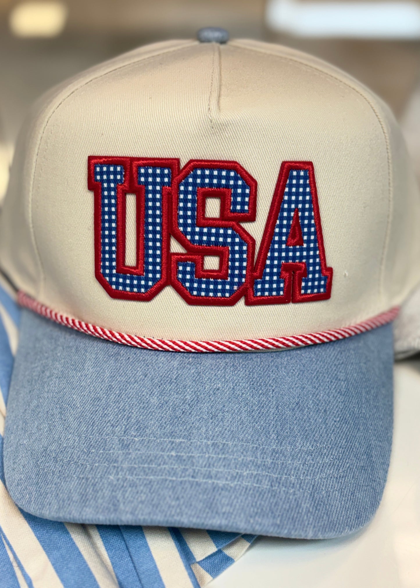Show your patriotic pride in this comfy cotton trucker hat with beige and blue gingham, bold USA letters, and striped rope trim. Elle Boutique, Zachary, LA.