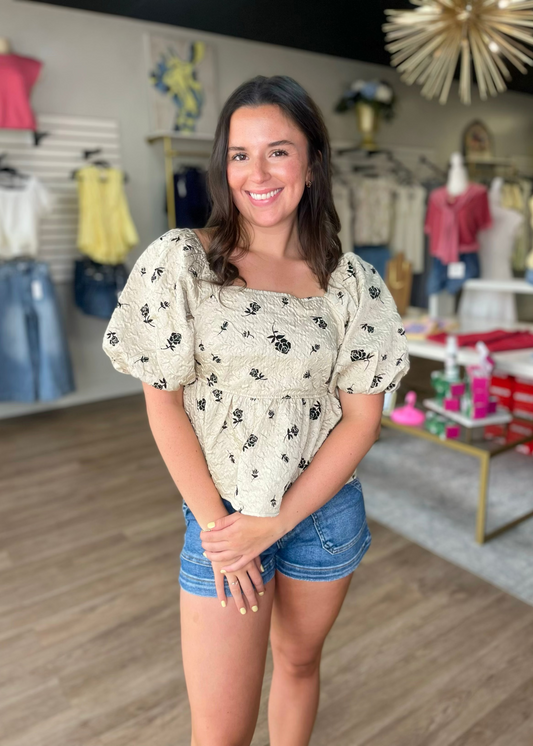 A woman grins in a blush pink floral peplum top with puff sleeves and comfy denim shorts. Elle Boutique, Zachary, LA.