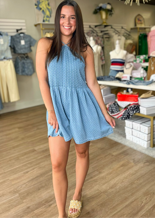 A woman beams in Elle Boutique, Zachary, LA, wearing a light blue denim romper with a relaxed fit and classic button details.