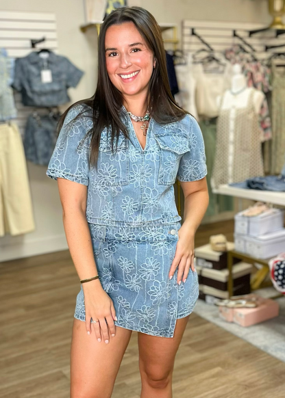 A woman with long brown hair beams in a soft denim, blue floral romper with a relaxed western fit at Elle Boutique, Zachary, LA.