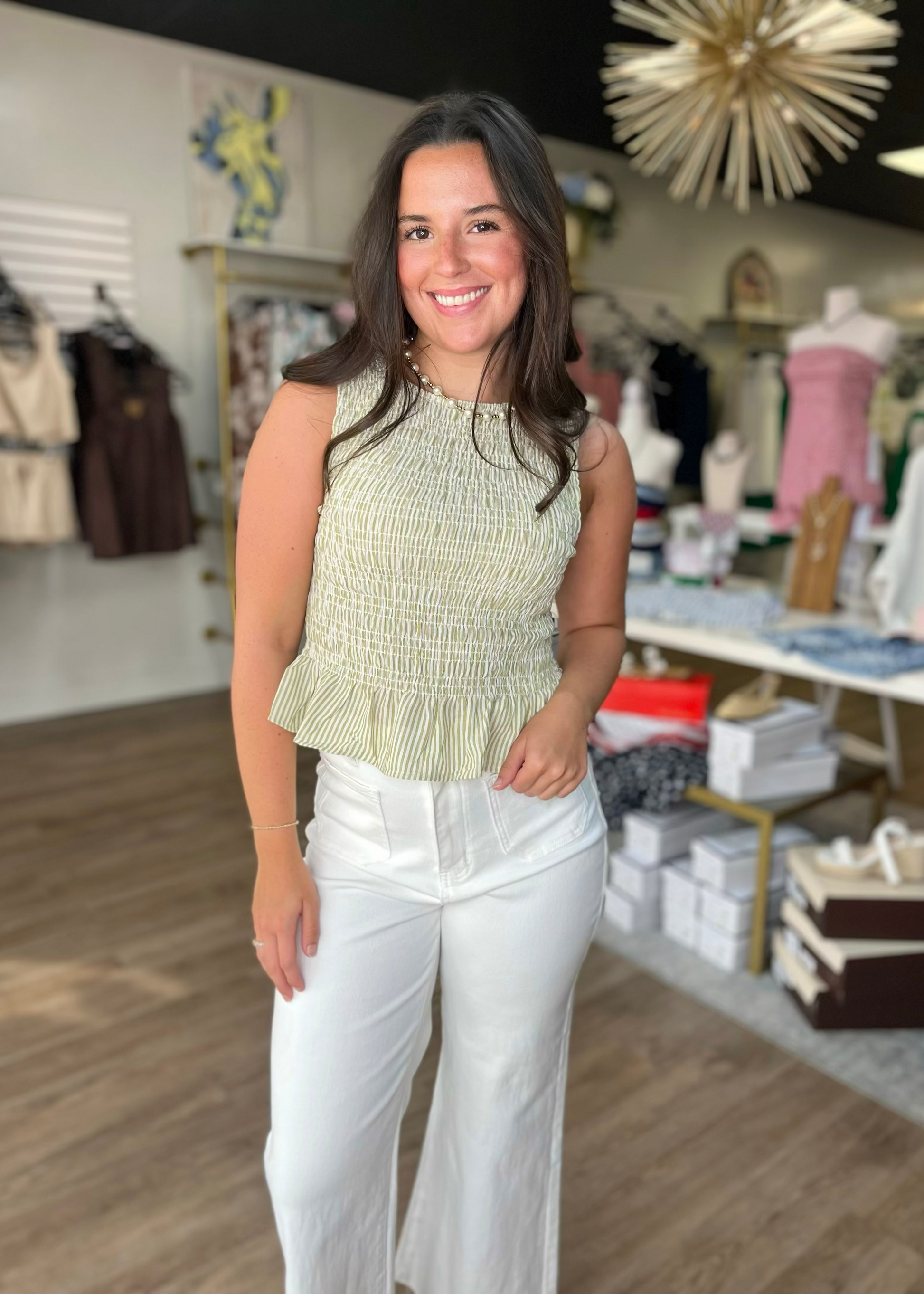 A cheerful woman in a soft olive smocked top and comfy white pants stands in a cozy shop. Elle Boutique, Zachary, LA.