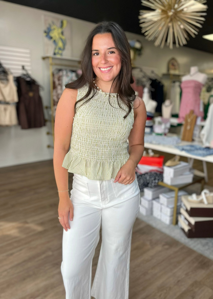 A cheerful woman in a soft olive smocked top and comfy white pants stands in a cozy shop. Elle Boutique, Zachary, LA.