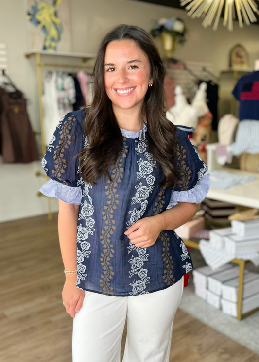 A woman in a soft navy cotton blouse with white floral embroidery and comfy white pants beams inside Elle Boutique, Zachary, LA.