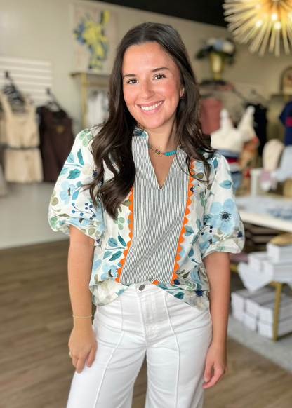 A smiling woman in a soft cotton Blue Blossom Scallop Trim Floral Top with white pants, inside Elle Boutique, Zachary, LA.