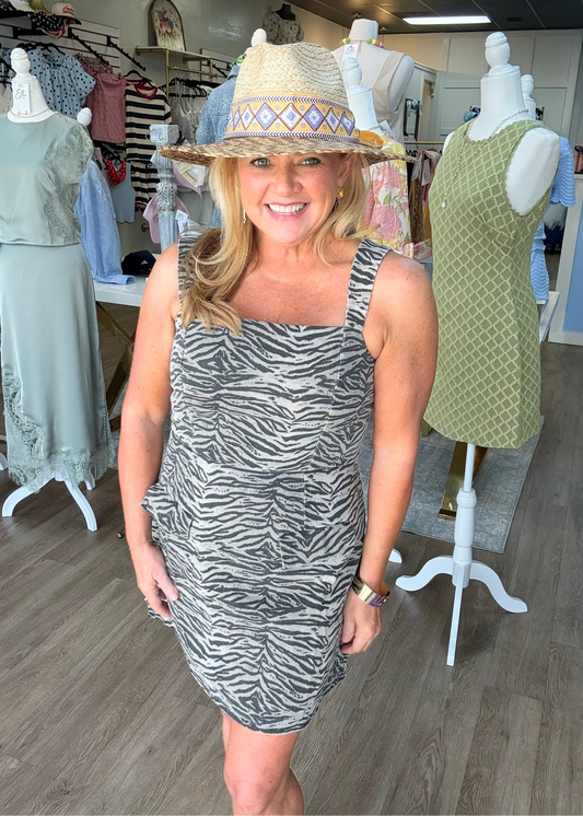 A cheerful woman with blonde hair stands on a wooden floor, wearing the Wild Side Tiger Print Square Neck Romper in soft, breezy fabric. The romper features a classic square neckline, comfy adjustable straps for the perfect fit, and bold tiger stripes in rich earth tones. She pairs it with a playful straw hat and has one hand tucked casually in her pocket. Around her, mannequins show off other darling styles inside a charming boutique setting. Elle Boutique, Zachary, LA.