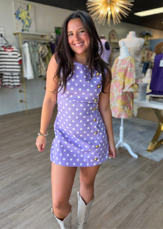 A lovely woman beams with Southern charm inside our boutique, standing below a whimsical star-shaped light and surrounded by delightful clothing racks. She wears the Scalloped Sweetheart Polka Dot Romper in a vintage-inspired lavender, dotted with crisp white polka dots and finished with gleaming gold buttons. The soft fabric gently hugs her frame, featuring a flattering sweetheart neckline and playful scalloped edges—perfect for porch gatherings or weekend strolls. Elle Boutique, Zachary, LA.