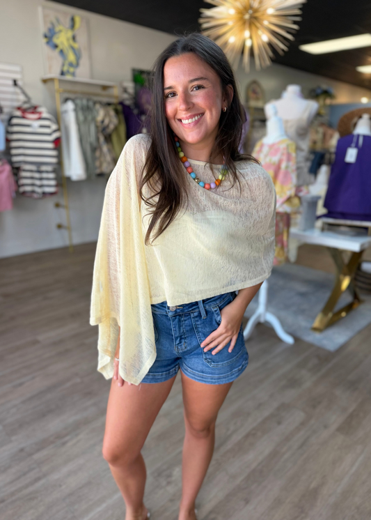 A cheerful woman is all smiles in the Golden Hour Sheer Knit Cape Shawl Poncho, draped light and airy over her soft yellow blouse. The gentle knit fabric flows with an easy, relaxed fit that pairs perfectly with her casual denim shorts and pops of color from a beaded necklace. She stands inside a cozy boutique filled with charming clothing racks, ready for sweet summer days. Elle Boutique, Zachary, LA.