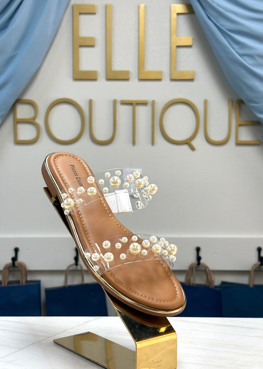 Pearl Luxe Clear Strap Sandal with shiny pearls and comfy clear straps rests on a gold stand by blue drapes. Elle Boutique, Zachary, LA.