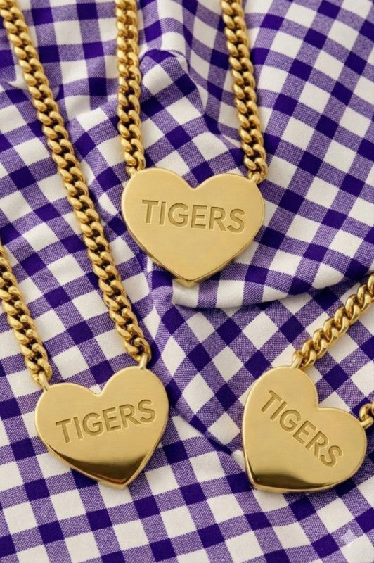 Three charming gold-tone Heart of a Tiger necklaces are displayed on a purple and white gingham fabric, perfect for showing your team spirit in sweet Southern style. Each heart-shaped pendant is carefully engraved with “TIGERS” and dangles from a bold, chunky gold chain—an adorable choice to layer over your favorite tee or dress on game day. Elle Boutique, Zachary, LA.